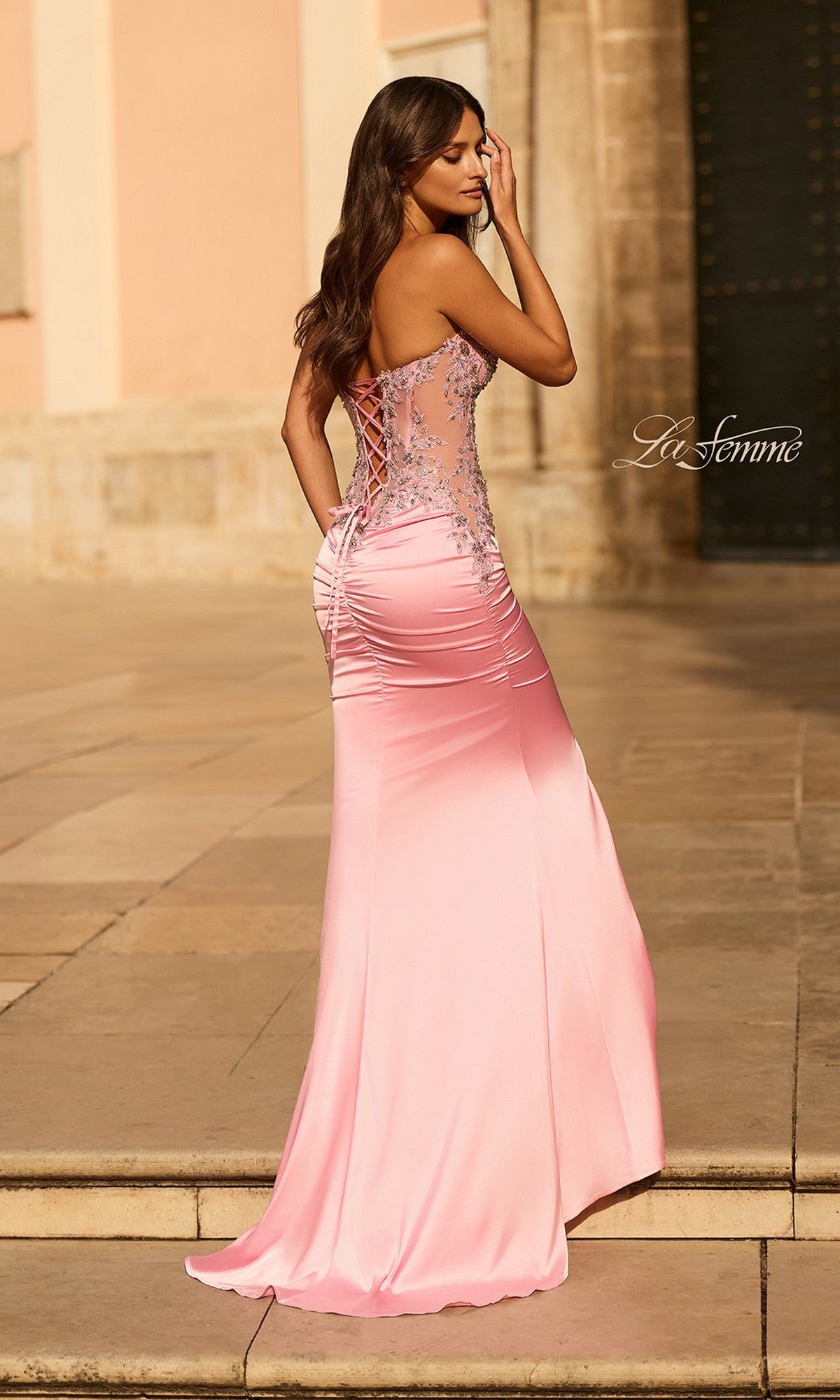 A woman stands outdoors in the La Femme 33765 long prom dress, featuring a strapless embellished corset bodice and lace-up open back, facing away from the camera.